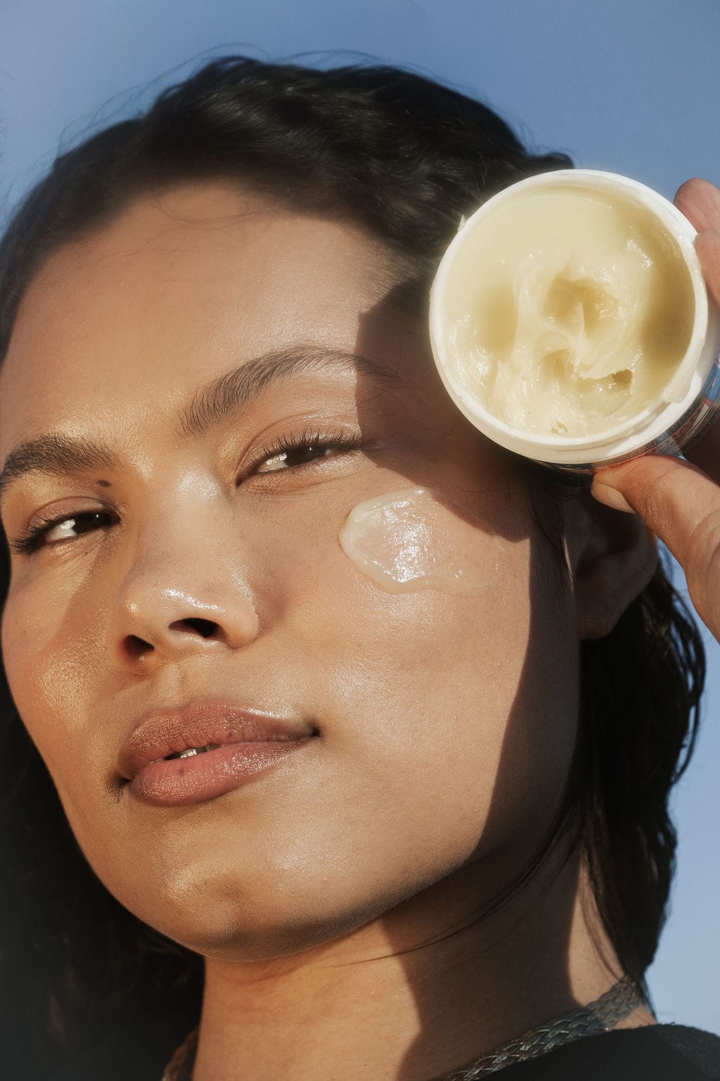 Model with an open jar of balm held beside her cheek, a swatch on her cheekbone