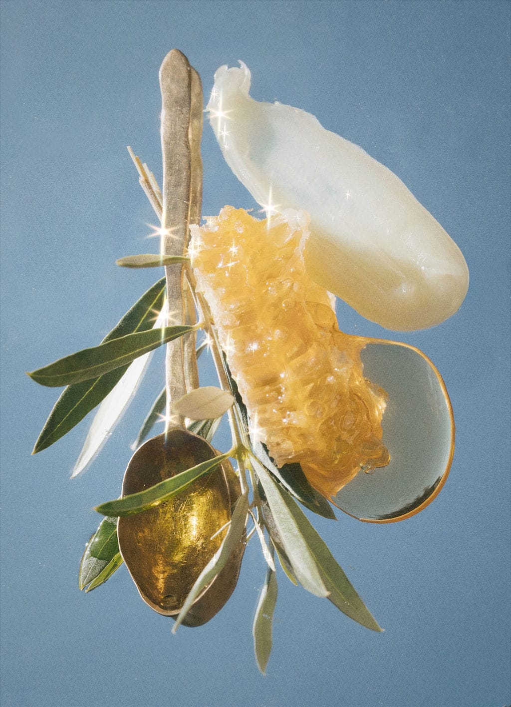 Olive branch, spoon of olive oil, raw honeycomb, balm and a honey drop suspended against slate blue