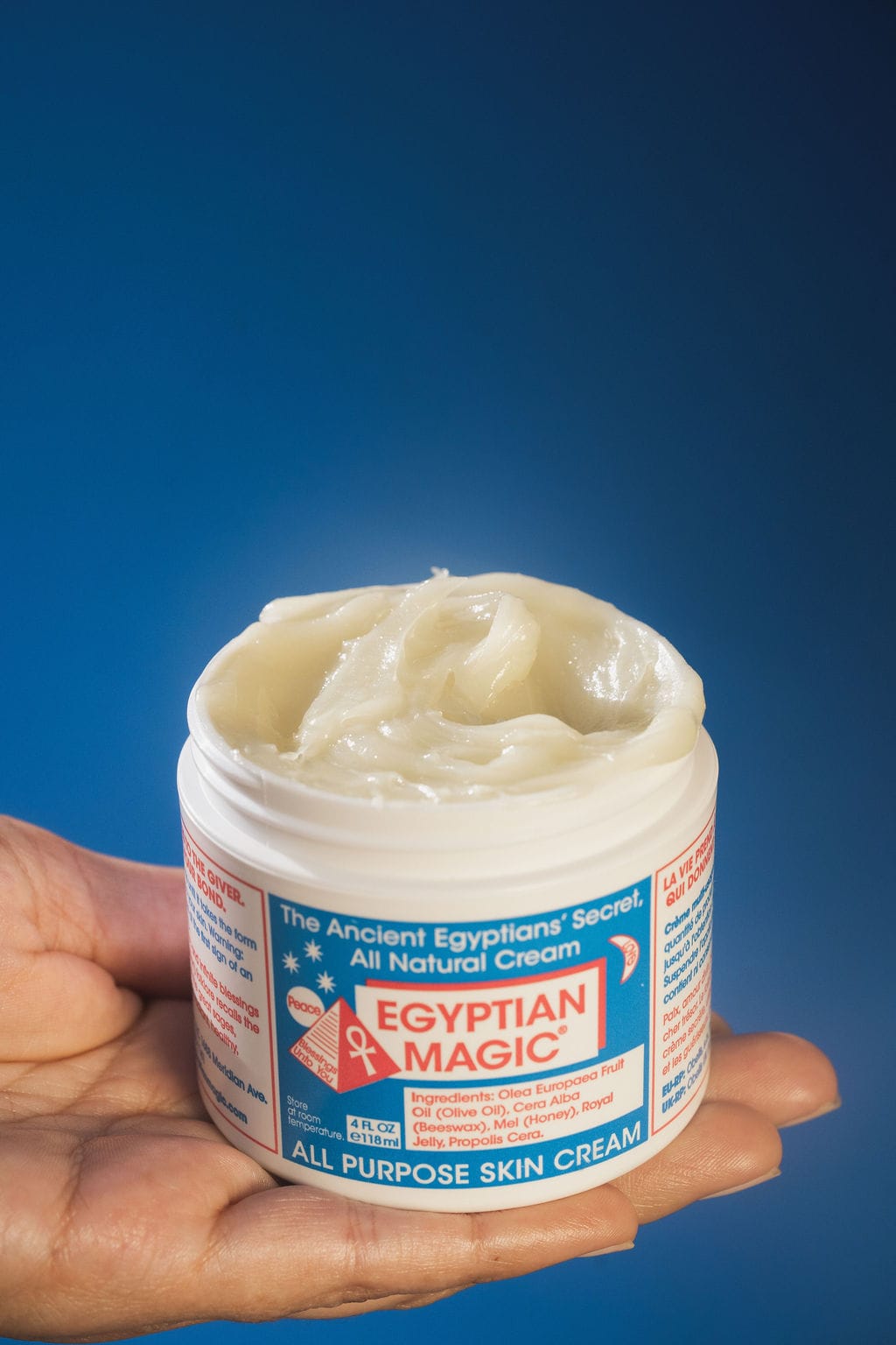 An open jar of Egyptian Magic held flat on a palm against a deep blue backdrop, the swirled balm texture visible